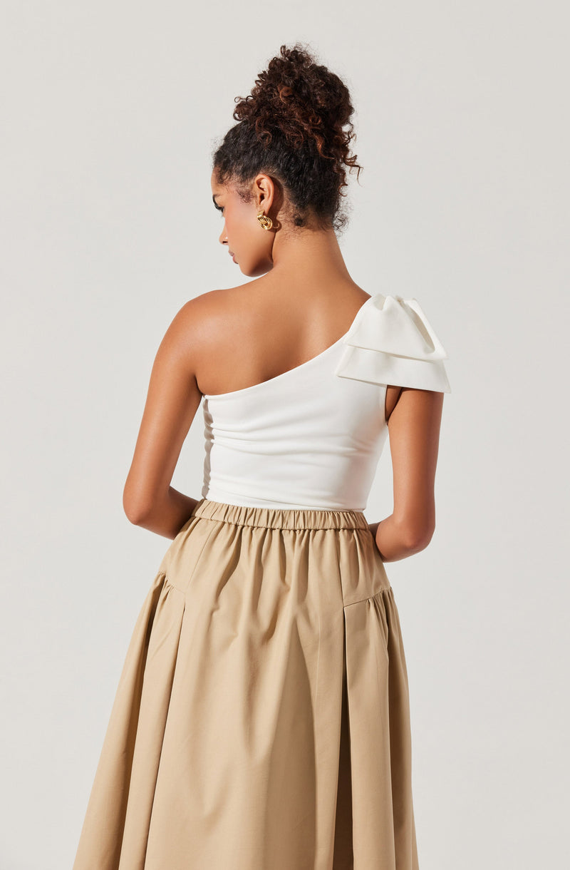 A woman is wearing a one-shoulder white top with a large, layered ruffle detail on the shoulder. She pairs it with a high-waisted, beige skirt that has pleats and an elastic waistband.