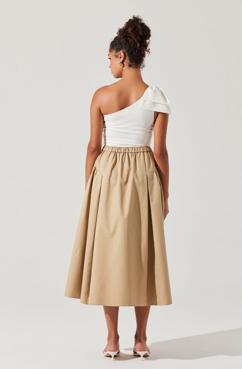 A woman is wearing a white one-shoulder top with a large bow detail on the shoulder, paired with a high-waisted, beige midi skirt that has a gathered elastic waistband and pleats. She is also wearing open-toed heels.