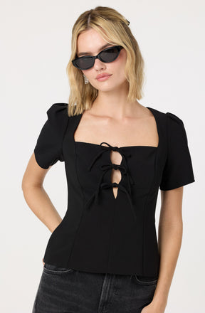 A woman is wearing a black, fitted short-sleeve top with puffed sleeves and a square neckline. The top features a distinctive front detail with three tied bows in the center. She has blonde, wavy hair and is accessorizing with large, black sunglasses and drop earrings.