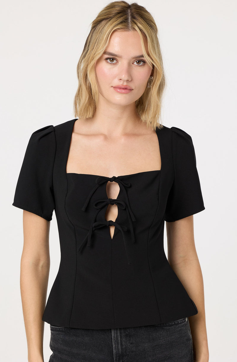A woman is wearing a black top with short, puffed sleeves and a square neckline. The top features three vertical cutouts in the front, each tied with a bow. The fabric appears smooth and fitted at the waist.
