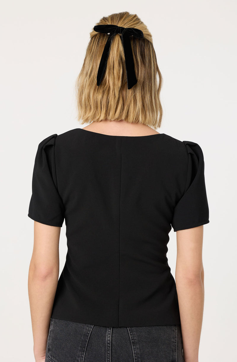 A woman is wearing a black short-sleeved top with puffed shoulders and a round neckline. She has a black ribbon tied into a bow in her blonde hair.