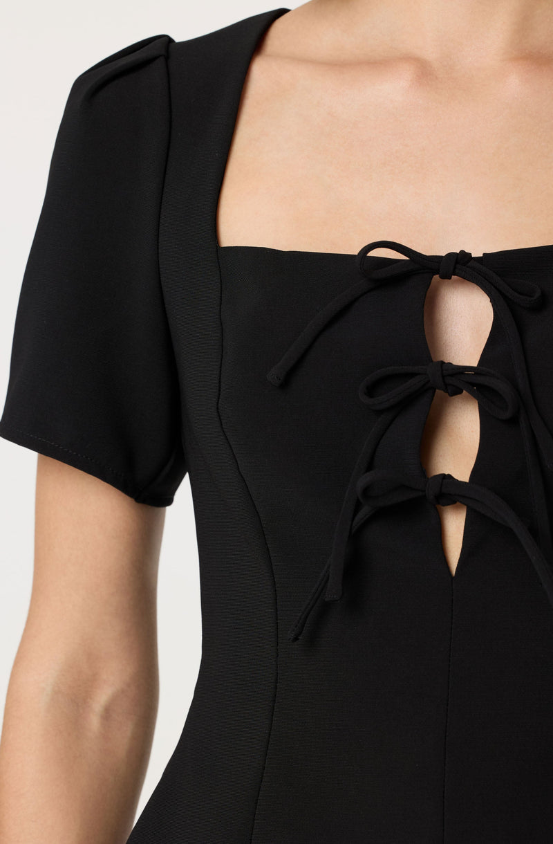 A close-up of a black dress featuring short puff sleeves and a square neckline with three decorative ties that create keyhole openings down the front.