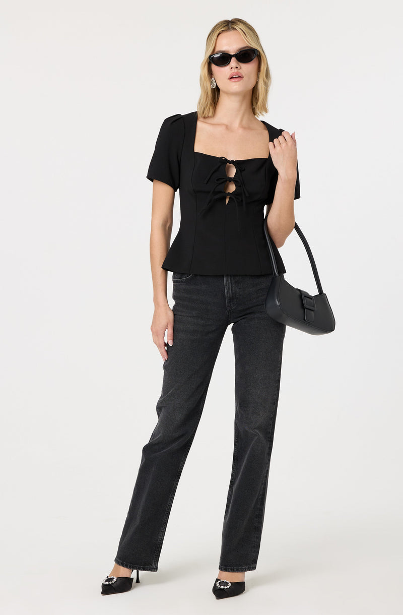 A woman is wearing a black top with short puff sleeves and a square neckline, featuring a lace-up detail with cutouts at the front. She pairs it with dark gray, straight-leg jeans and black pointed-toe heels with embellishments. She accessorizes with black sunglasses, earrings, and carries a black shoulder bag with a rounded shape.