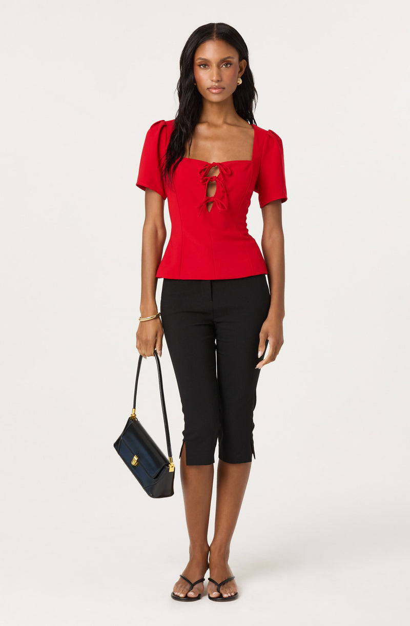 A woman is wearing a red top with short puff sleeves and a square neckline featuring a lace-up detail in the front. She pairs it with black knee-length pants that have small slits at the hem. She is accessorizing with a black handbag with gold accents, gold hoop earrings, and a bracelet, and is wearing black strappy sandals.