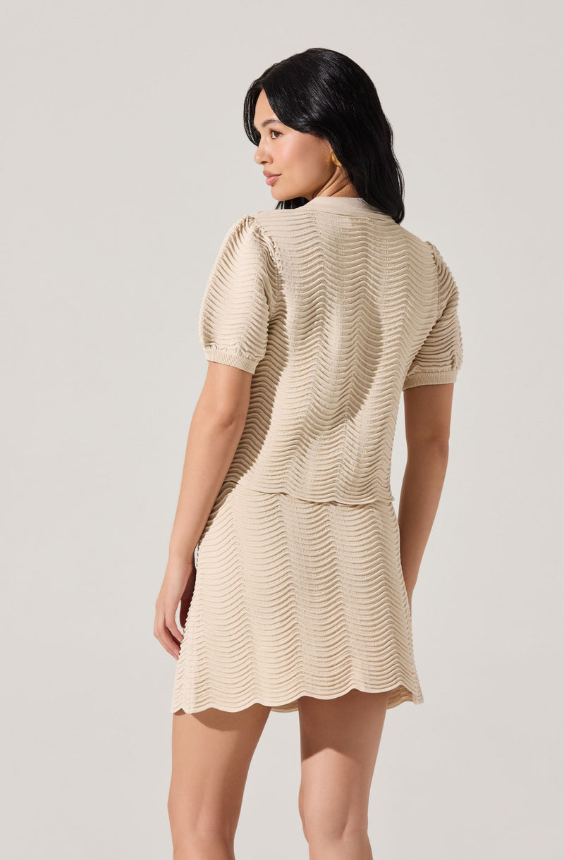 A woman is wearing a beige knit dress with textured horizontal wavy stripes. The dress has short puffed sleeves and a round neckline. The hem of the dress is above the knee, and the fabric appears soft and slightly stretchy.