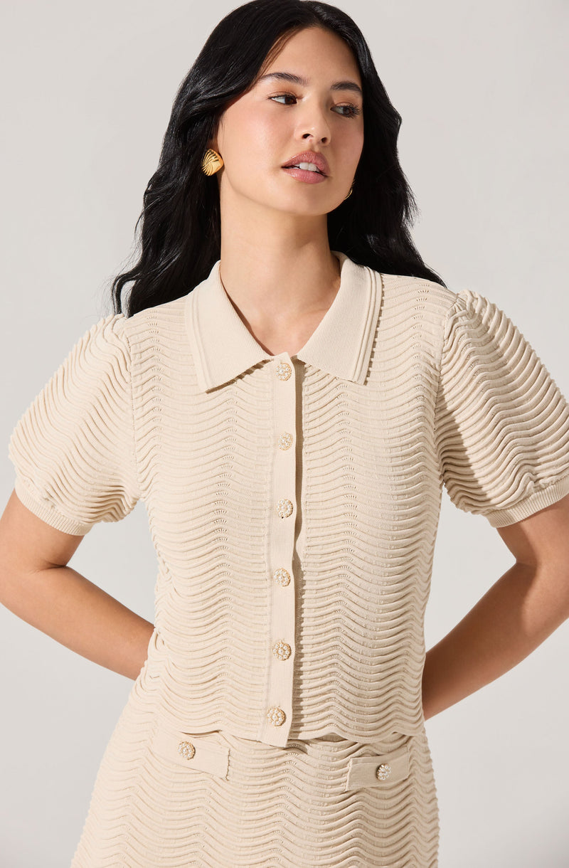 A woman is wearing a cream-colored, textured short-sleeve button-up top with a collar. The top has a ribbed, wavy pattern throughout and decorative buttons along the front. She is also wearing matching textured bottoms with button details at the waistband. She has black, wavy hair and gold earrings.