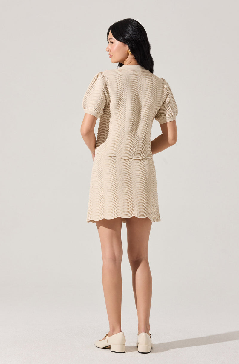 A woman is wearing a matching beige outfit consisting of a textured, short-sleeved top and a matching textured skirt. The top has a subtle wave pattern, puffed sleeves, and a round neckline. The skirt is above the knee with a similar wave pattern and a scalloped hem. She is also wearing off-white block-heeled shoes and gold earrings.
