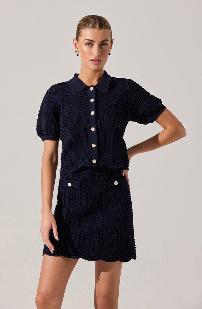 A woman is wearing a matching navy blue textured two-piece outfit consisting of a short-sleeved, collared shirt and a skirt. The shirt features a row of white buttons down the front, and the skirt has a similar textured pattern with decorative white buttons on the front pockets. The fabric has a horizontal ribbed or wave-like pattern, giving it a distinctive textured appearance.