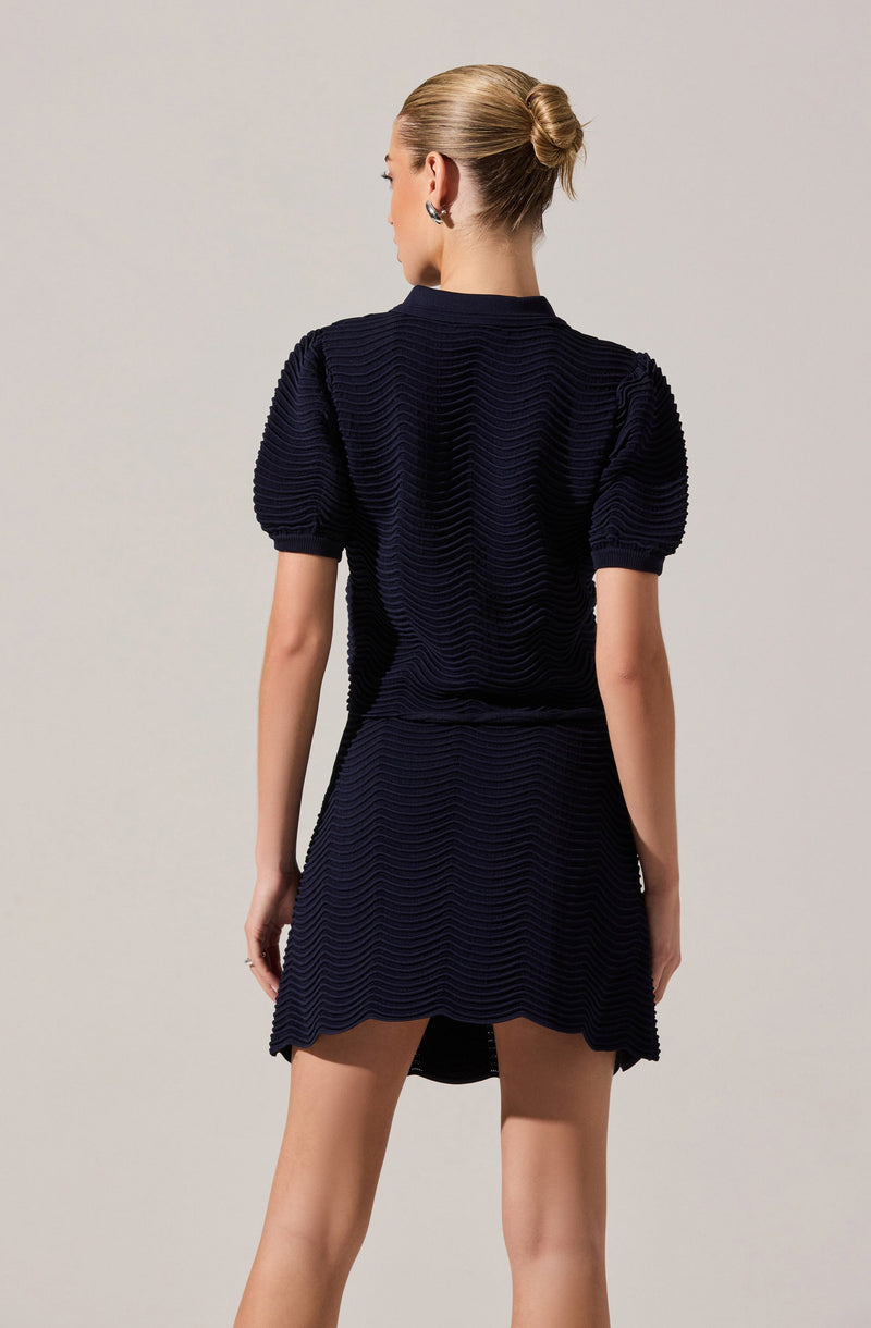 A woman is wearing a matching set consisting of a navy blue textured top with puffed short sleeves and a matching skirt. The top has a ribbed, wave-like pattern, and the skirt features the same textured pattern with a slightly flared hem. She has her hair styled in a sleek, low bun and is wearing earrings.