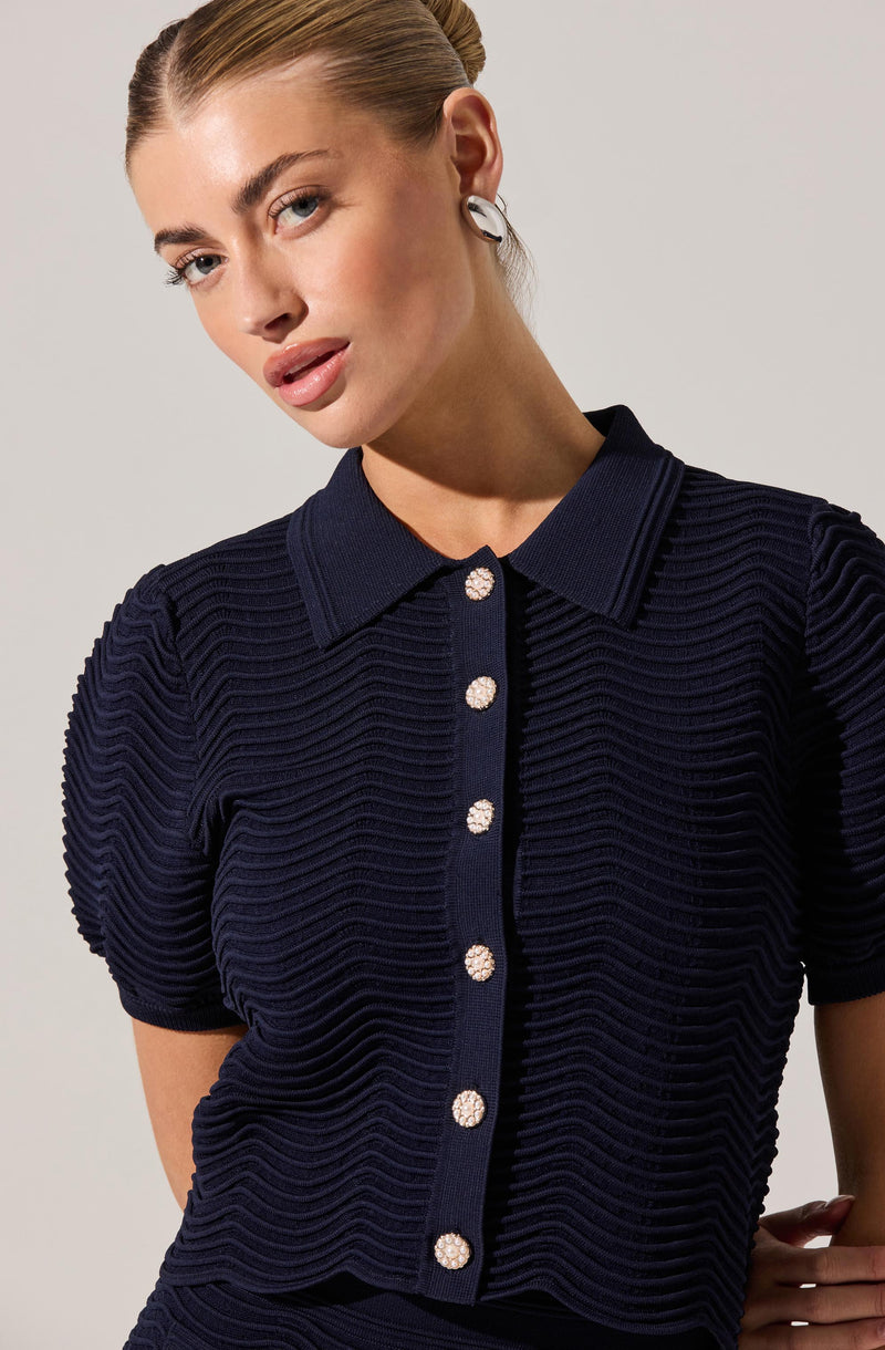 A woman is wearing a navy blue textured short-sleeve button-up top with a collar. The top features a ribbed, wavy pattern across the fabric, with decorative shiny, pearl-like buttons down the front.