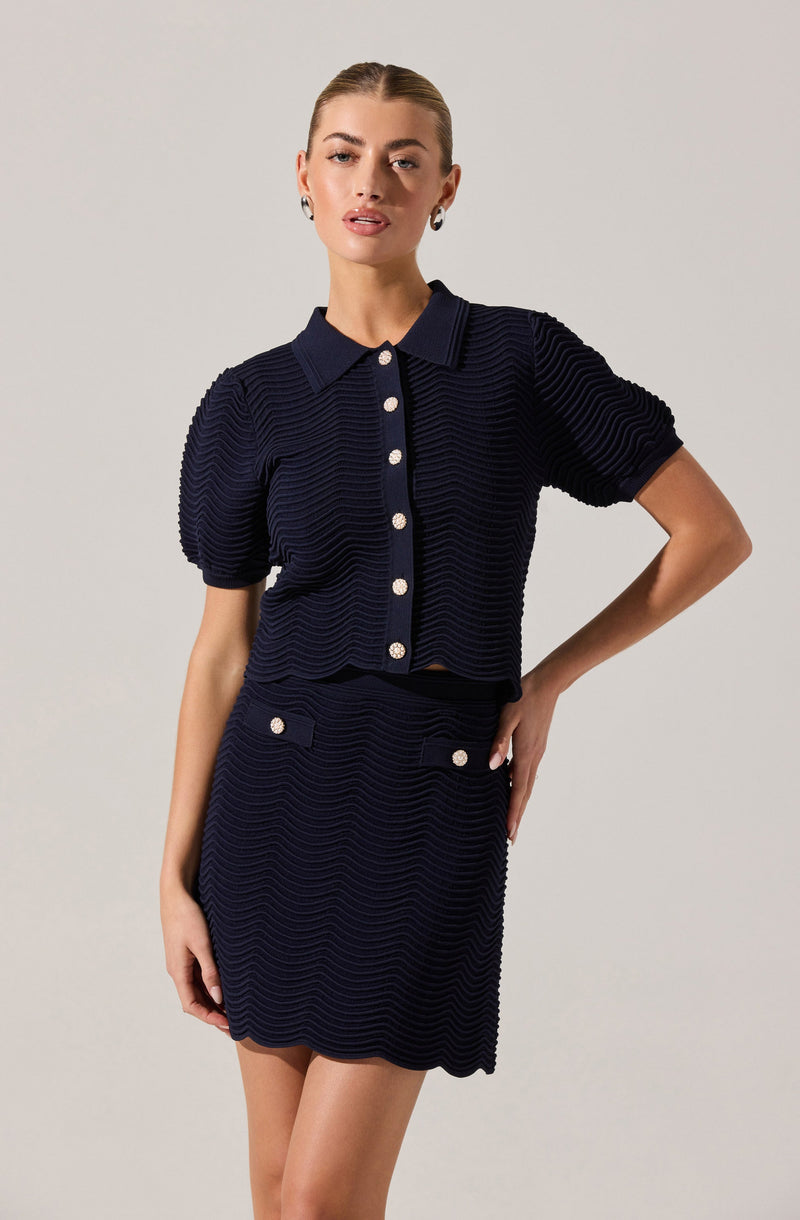 A woman is wearing a coordinated navy blue two-piece outfit consisting of a short-sleeved, collared button-up top and a matching skirt. The top features textured horizontal stripes and large decorative buttons down the front. The skirt, also textured with horizontal stripes, has a high waist with decorative buttons on the waistband and a simple, straight silhouette.