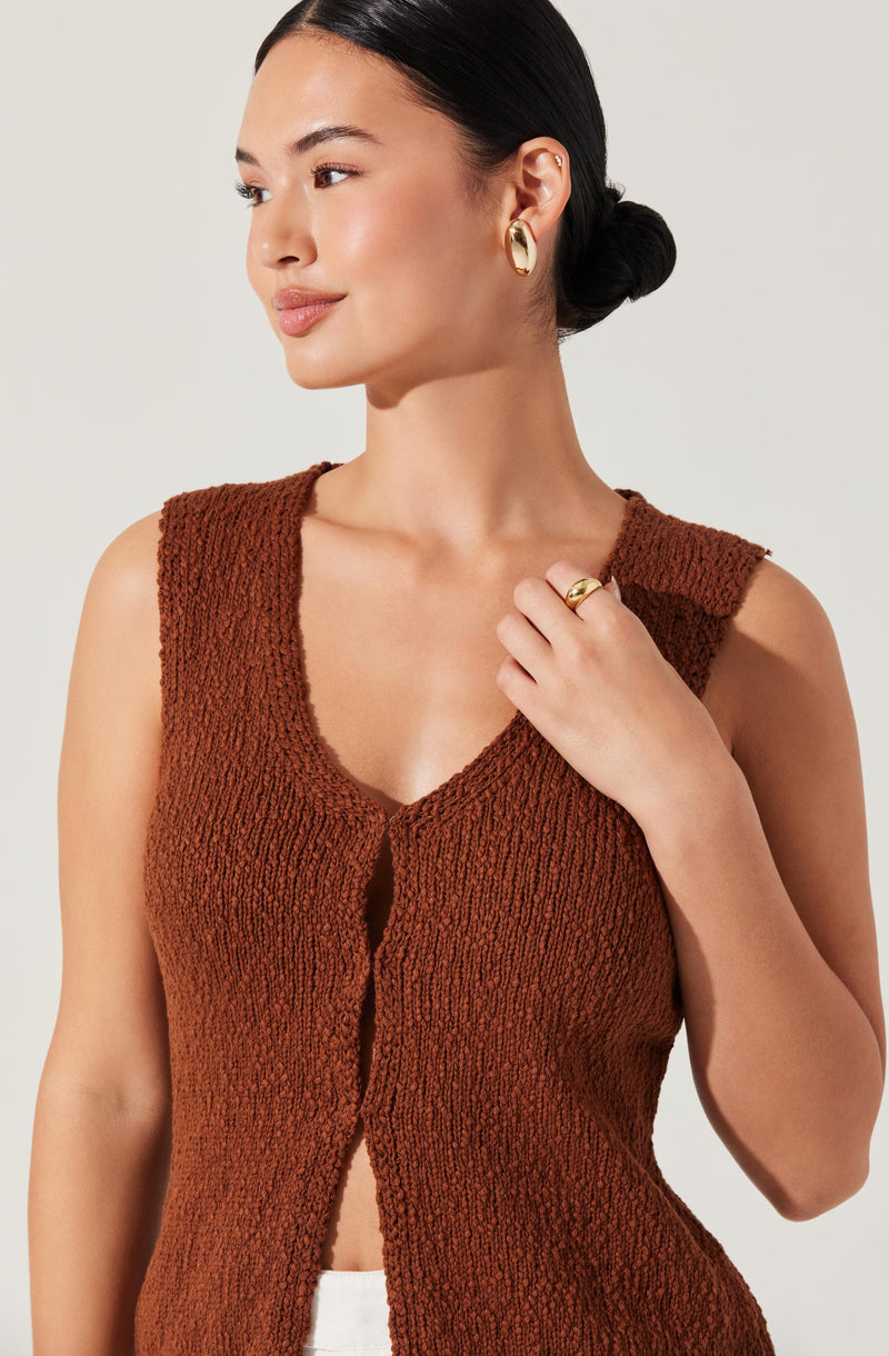 A woman wearing a sleeveless, rust-colored, textured knit top with a deep V neckline and a front opening. She has dark hair styled in a low bun, and she is accessorized with gold hoop earrings and a gold ring on her right hand.