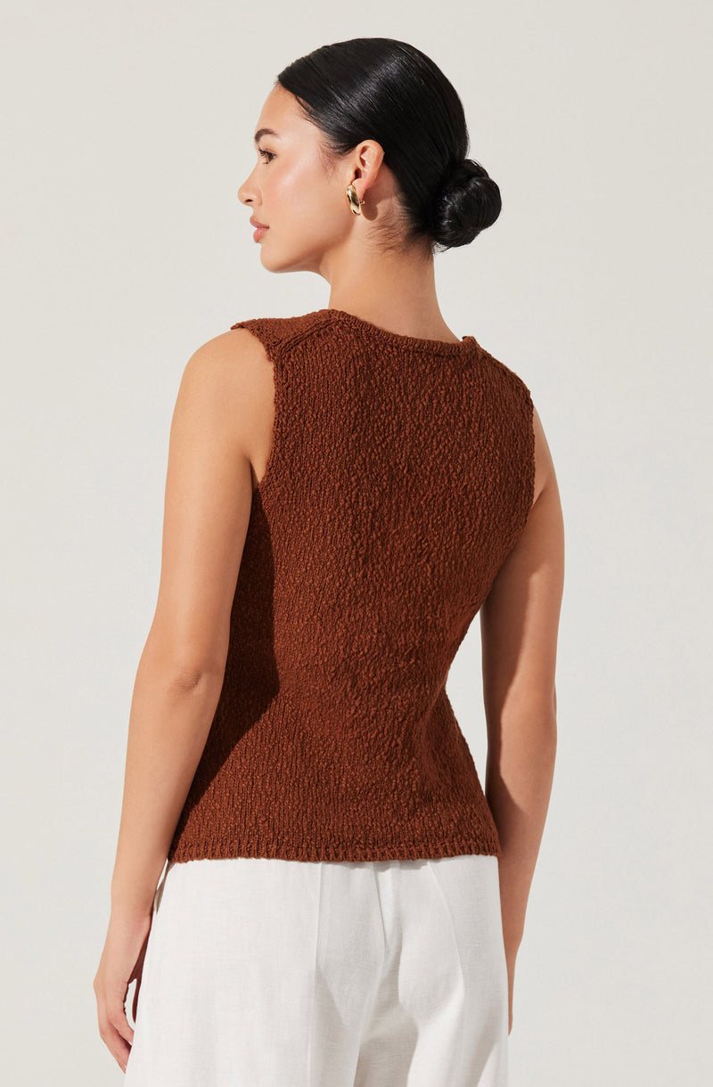 A woman is wearing a sleeveless, textured brown top that fits snugly at the waist. The top has a round neckline and a slightly raised armhole design. She pairs it with light-colored, high-waisted pants.