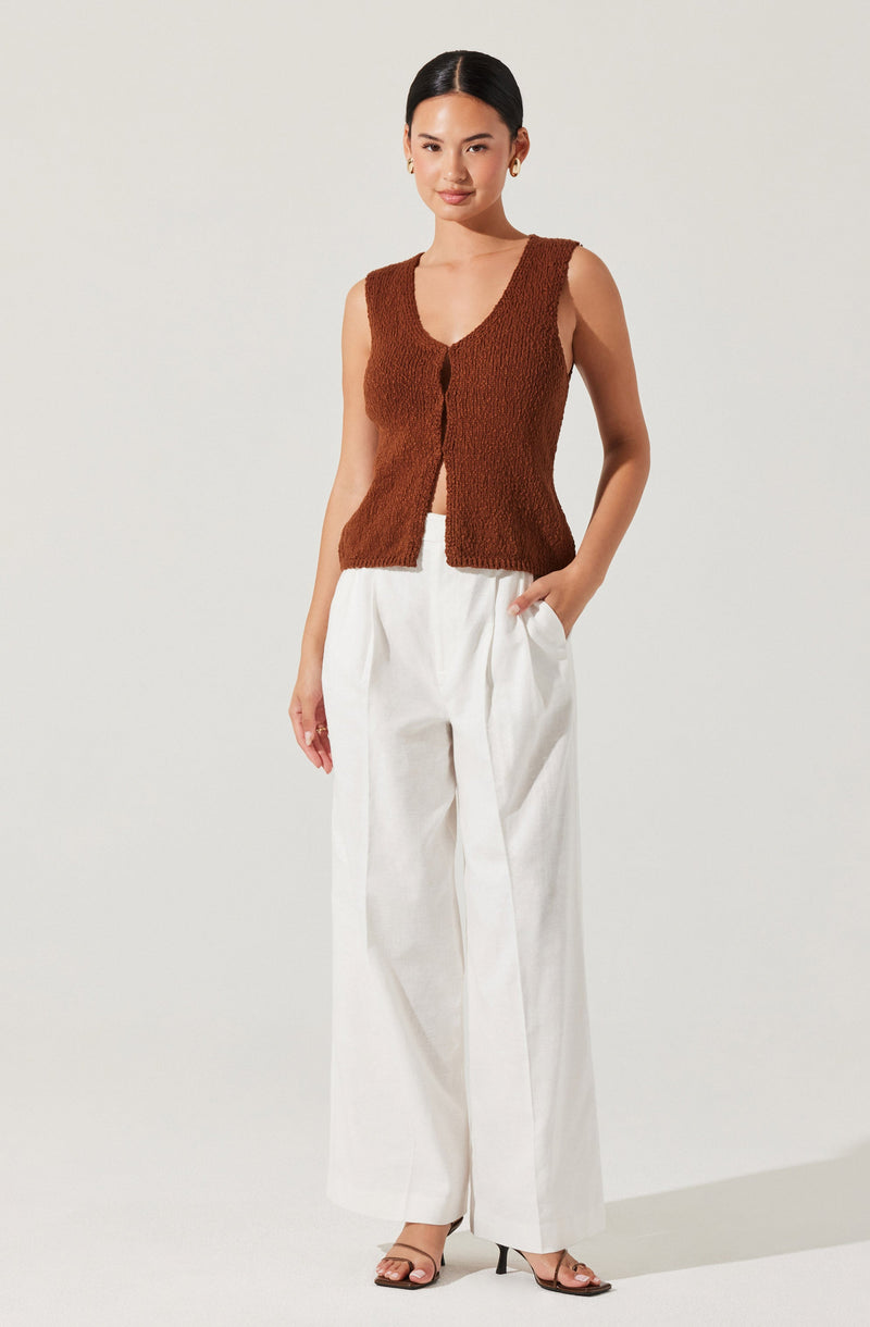 A woman is wearing a sleeveless, textured brown vest with a V-neckline and a button closure at the front. She pairs the vest with high-waisted, wide-legged white pants and strappy open-toe heels.