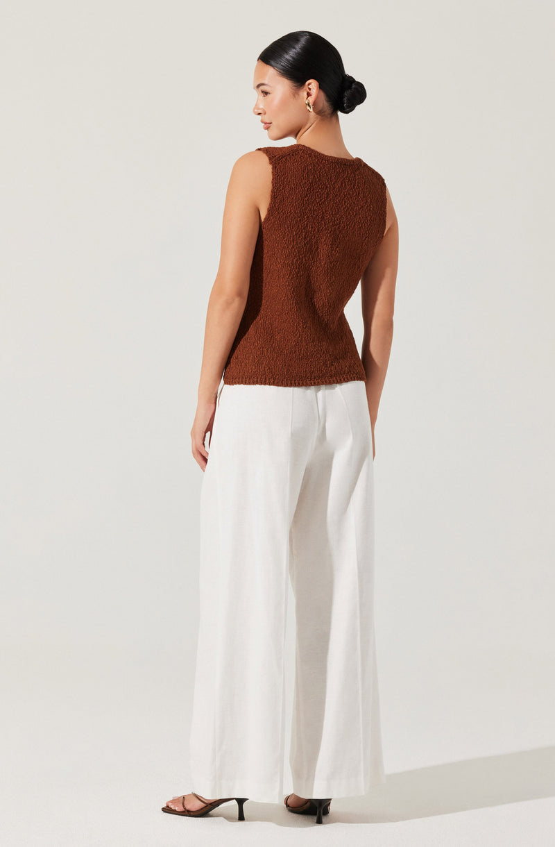 A woman is wearing a sleeveless, textured brown top with a high neckline and a slightly loose fit. She pairs it with wide-leg, high-waisted white pants that reach the floor, and is wearing black open-toe high heels. Her hair is styled in a neat, low bun.