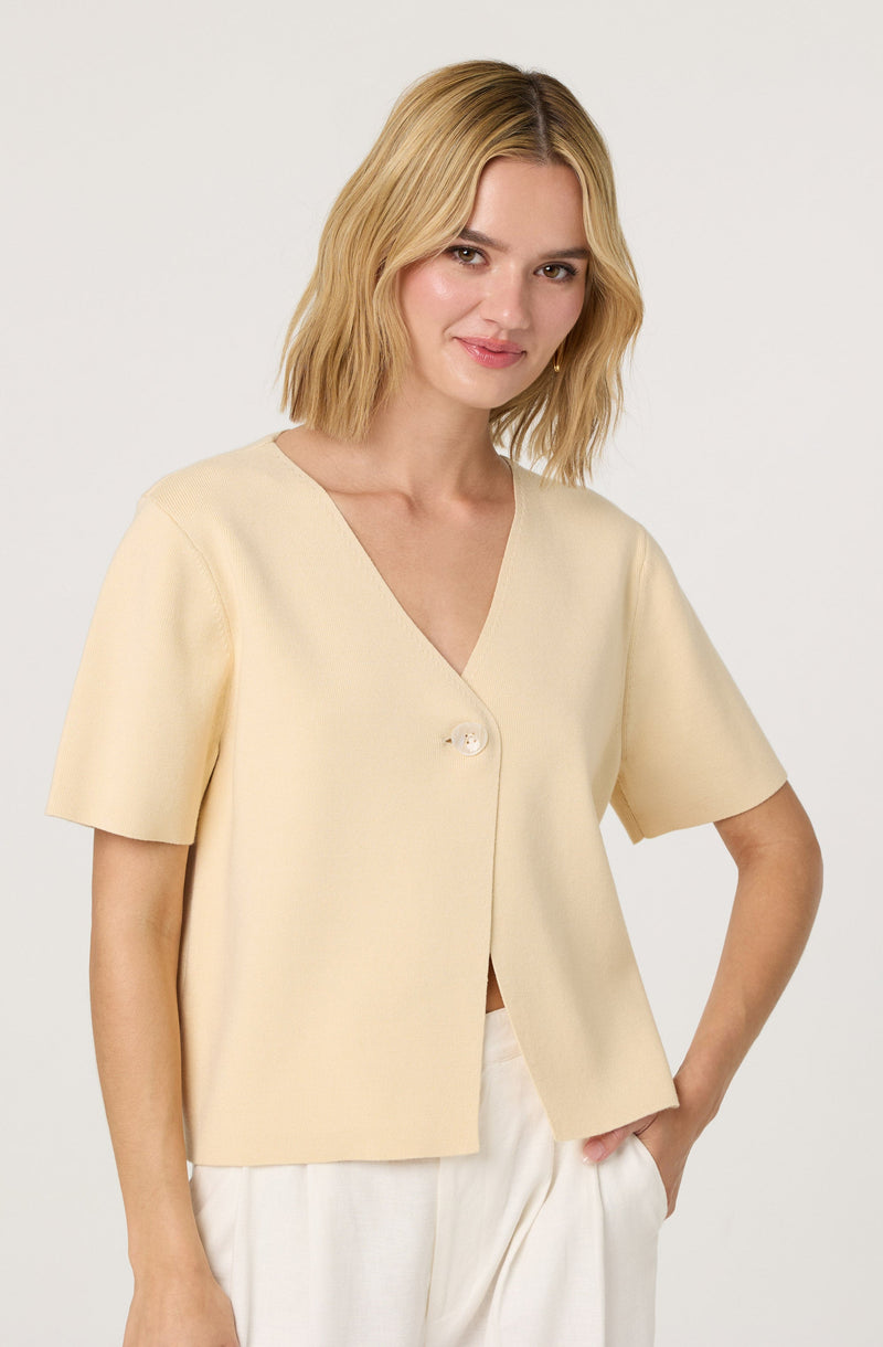 A woman is wearing a short-sleeved, cream-colored top with a V-neckline and a single large button closure near the chest. The top has a relaxed fit and appears to be made of a soft, textured fabric.