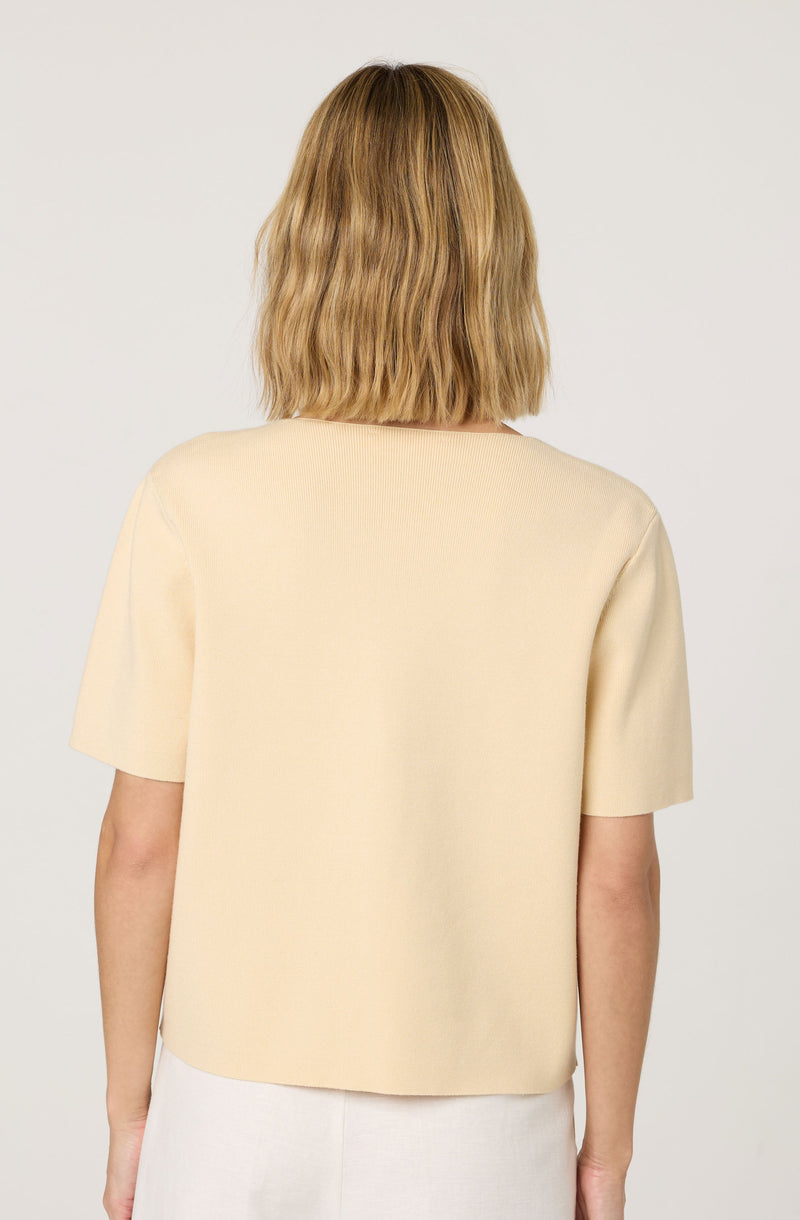 A woman is wearing a short-sleeved, beige top made of a textured, knit-like fabric. The top has a simple, round neckline and a loose fit. She is also wearing a light-colored skirt.