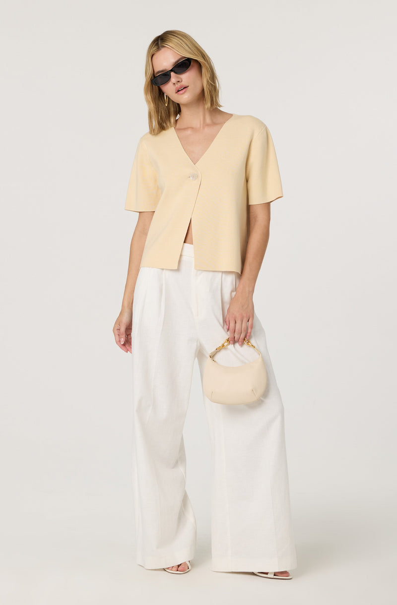 A woman is wearing a short-sleeved, light beige V-neck top with an asymmetrical button closure. She has on high-waisted, wide-leg white pants and is accessorizing with black sunglasses and a small, cream-colored handbag with a gold handle.