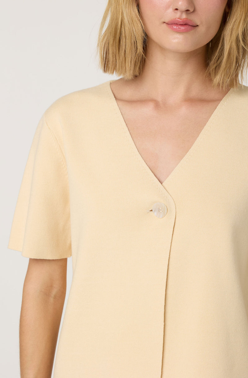 A woman is wearing a light beige, short-sleeved top with a V-neckline. The top features a single large, round, clear button near the center of the chest. The fabric appears to be soft and textured.