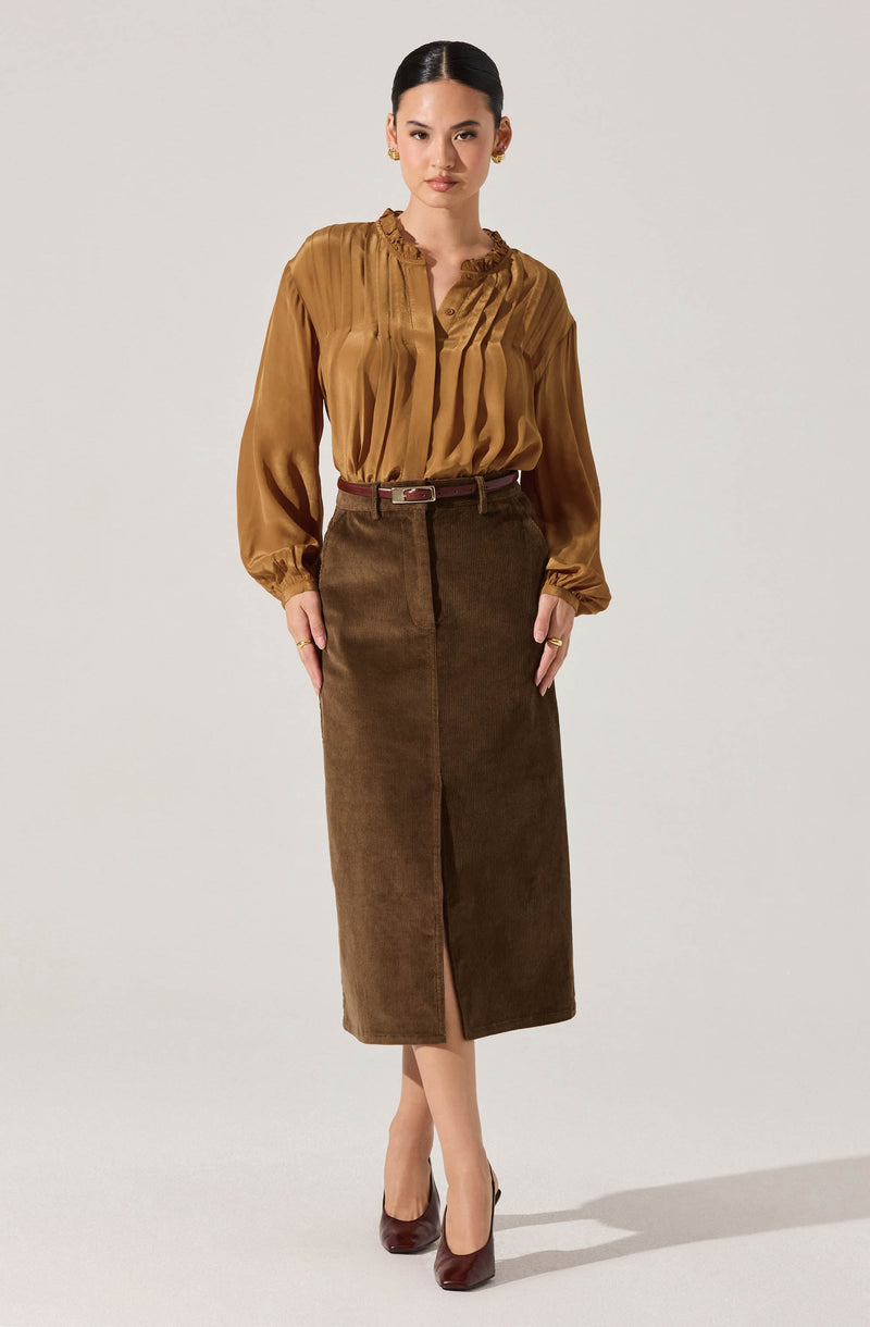 A woman is wearing a long-sleeved, shiny, satin-like blouse in a warm caramel color with pleated details on the front and ruffled trim around the collar. She pairs it with a high-waisted, brown corduroy midi skirt with a front slit, a belt in a matching brown shade, and dark burgundy high heels.