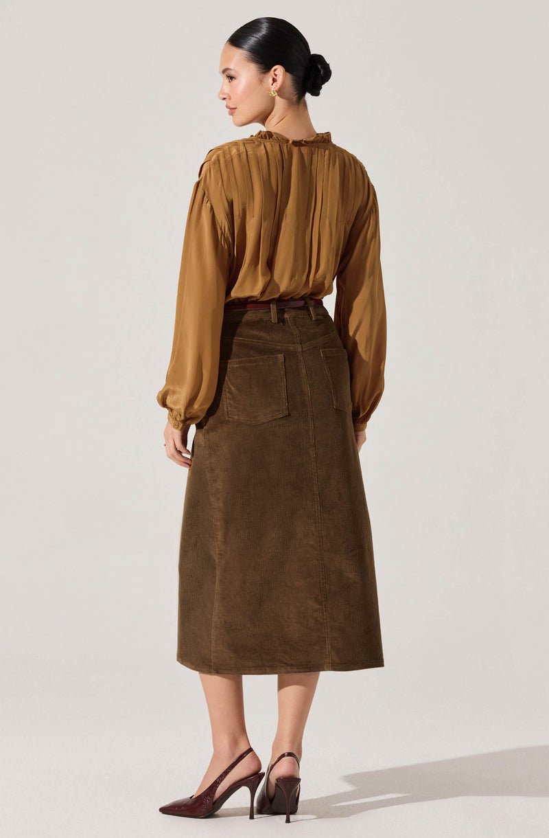 A woman is wearing a long-sleeved, mustard-colored blouse with a pleated design and a high neckline. She pairs it with a high-waisted, dark brown corduroy A-line skirt that reaches mid-calf, featuring large back pockets. She completes the outfit with burgundy high heel shoes.