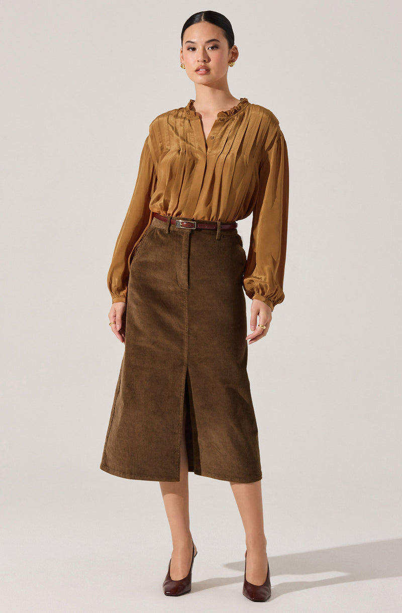 A woman is wearing a long-sleeved, satin blouse in a warm caramel color with ruffled details at the neckline and elasticized cuffs. She pairs it with a high-waisted, brown suede midi skirt featuring a front slit and a matching belt. She completes the look with dark brown high heels.