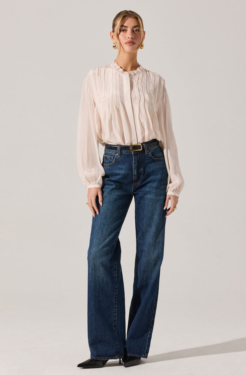 A woman is wearing a long-sleeved, light-colored blouse with vertical pleats and a ruffled neckline. She pairs it with high-waisted, dark blue wide-leg jeans that have a black belt with a gold buckle. She completes the outfit with black pointed shoes.