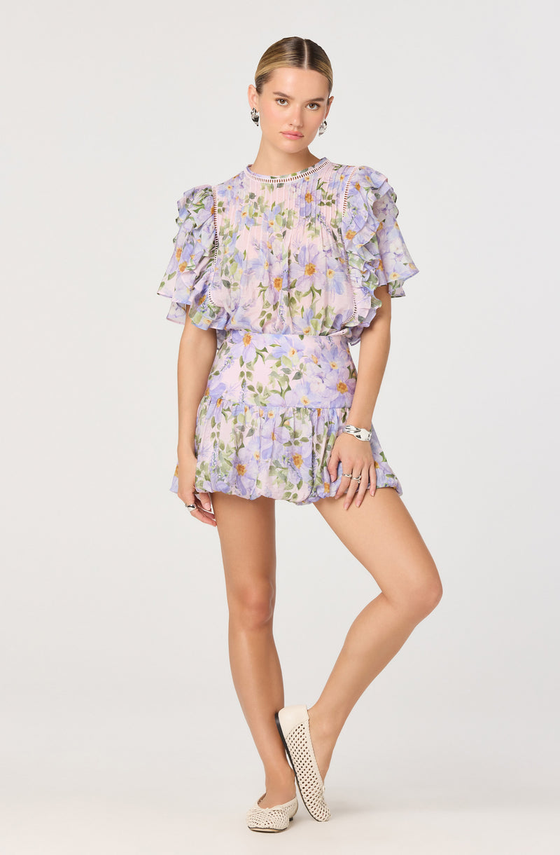 A woman is wearing a two-piece outfit with a pastel-colored floral print. The top has ruffled short sleeves with layered ruffles and detailed trim along the edges. The matching skirt is short, with a fitted waist and a ruffle hem that complements the top's ruffles.