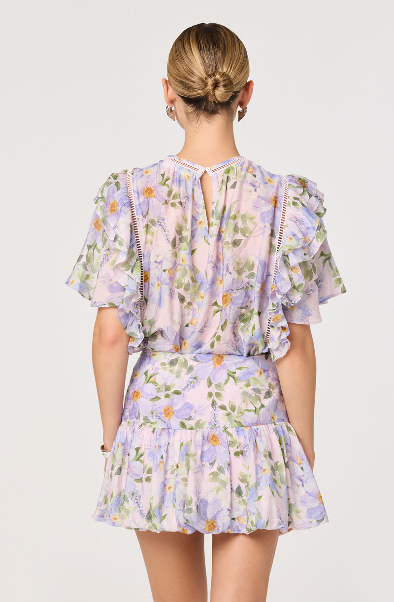 The outfit consists of a matching romper with a pastel floral print in shades of lavender, yellow, green, and white. The top has ruffled short sleeves with decorative trim along the edges and a small keyhole opening at the back. The shorts are high-waisted with a gathered, ruffled hem, creating a layered appearance. The fabric appears light and airy, suitable for warm weather.