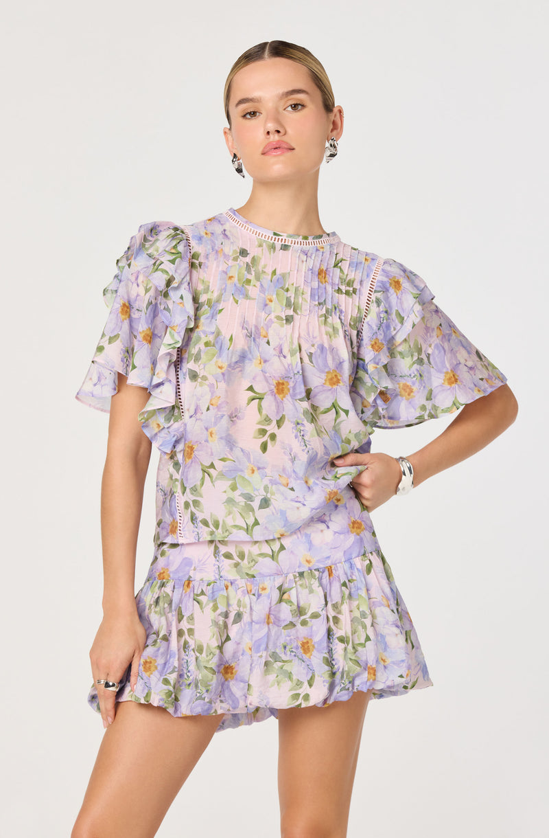 A woman is wearing a matching two-piece outfit consisting of a short-sleeved top and a skirt with a floral pattern. The top has ruffled sleeves and a round neckline with lace trim, made from a light, semi-sheer fabric with purple, green, yellow, and white flowers. The skirt is a ruffled mini-length, coordinating with the top's floral pattern and colors.