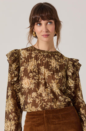 A woman is wearing a brown long-sleeve blouse with a floral print pattern in shades of beige and cream. The blouse features ruffled details along the shoulders and a gathered neckline. She is also wearing a high-waisted, brown corduroy skirt. She has gold hoop earrings and shoulder-length brown hair with bangs.