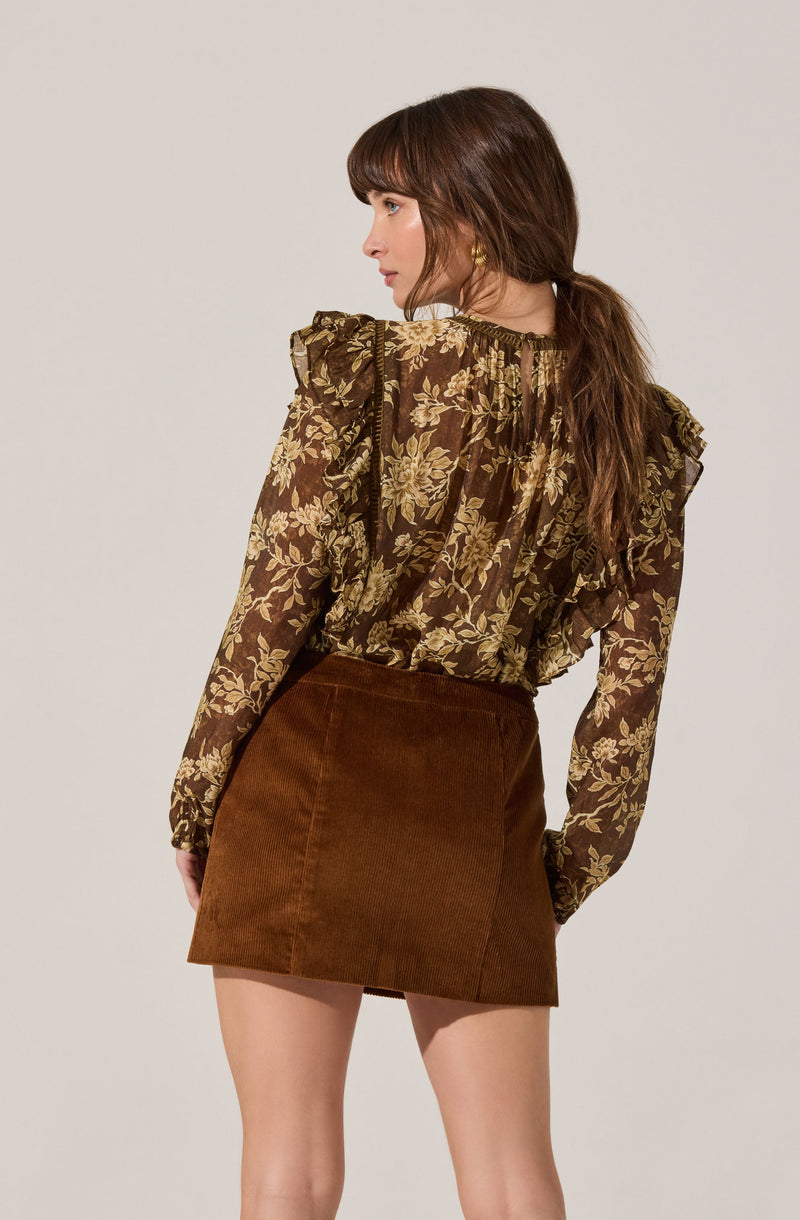 A woman is wearing a sheer, long-sleeved blouse with a brown background and a gold floral pattern. The blouse has ruffled details on the shoulders and is slightly gathered at the neckline. She pairs it with a high-waisted, brown corduroy mini skirt. She has shoulder-length, wavy brown hair with bangs and is wearing small gold hoop earrings.