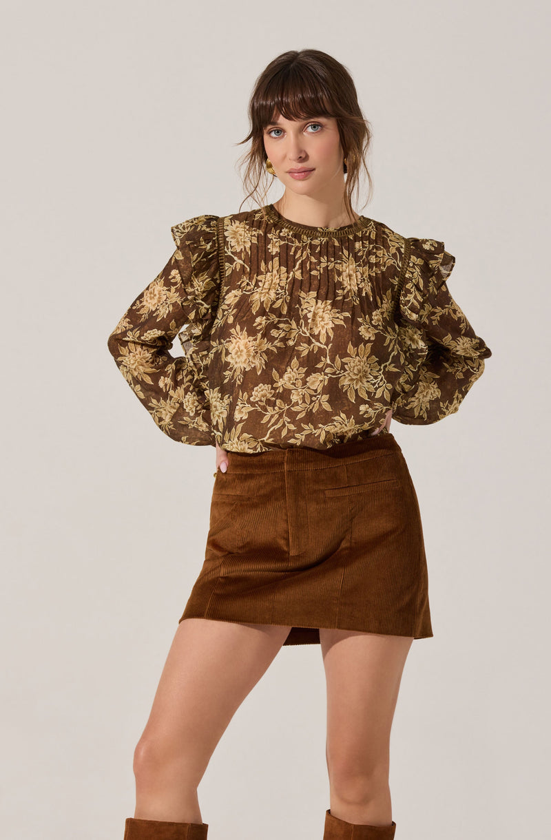 A woman is wearing a long-sleeved blouse with a dark brown background and a large, light beige floral pattern. The blouse has ruffled shoulder details and a gathered neckline. She pairs it with a high-waisted, brown corduroy mini skirt that has a front slit and patch pockets.