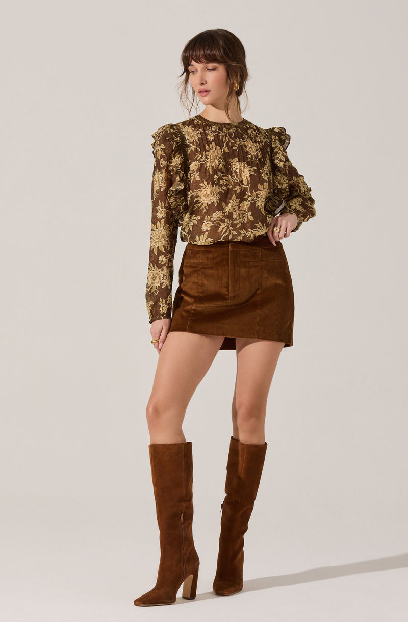 A woman is wearing a long-sleeved blouse with a floral pattern in shades of gold and brown, featuring ruffled shoulders. She pairs it with a brown suede mini skirt and matching knee-high suede boots with high heels.