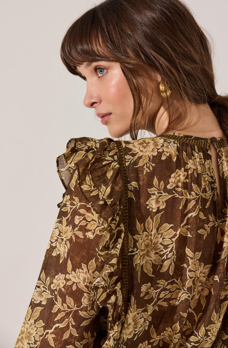 The woman is wearing a brown sheer blouse with a floral pattern in shades of beige and cream. The blouse has puffed sleeves with ruffles and is fastened at the back with a button closure at the neckline. She is also wearing gold earrings.