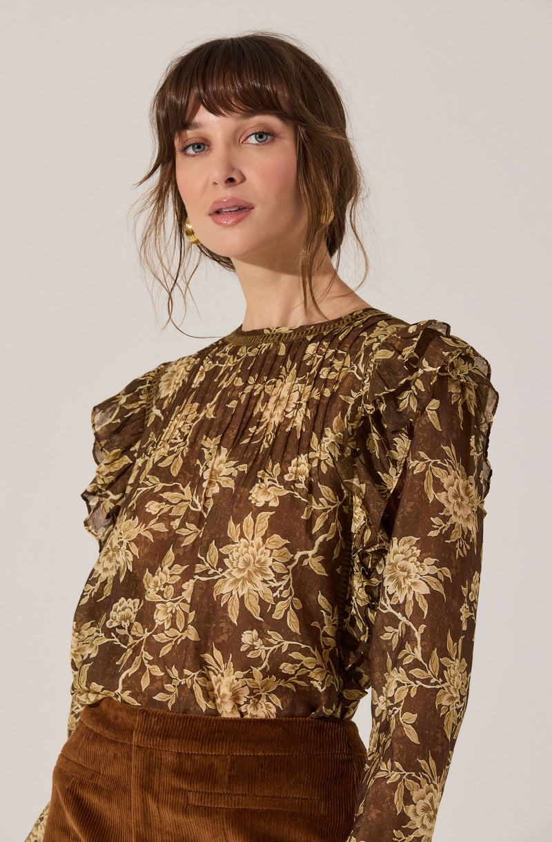 A woman is wearing a long-sleeved blouse with a brown background and a beige floral pattern. The blouse has pleated details on the yoke and ruffled shoulders, creating a textured and feminine look.