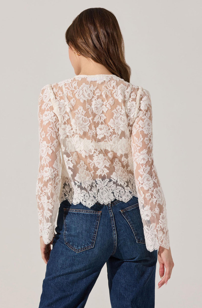 A woman is wearing a long-sleeved, sheer lace top with a floral pattern. The top is white or cream-colored, and the floral lace design is intricate, covering the entire back and sleeves. She pairs the top with high-waisted, dark blue jeans.