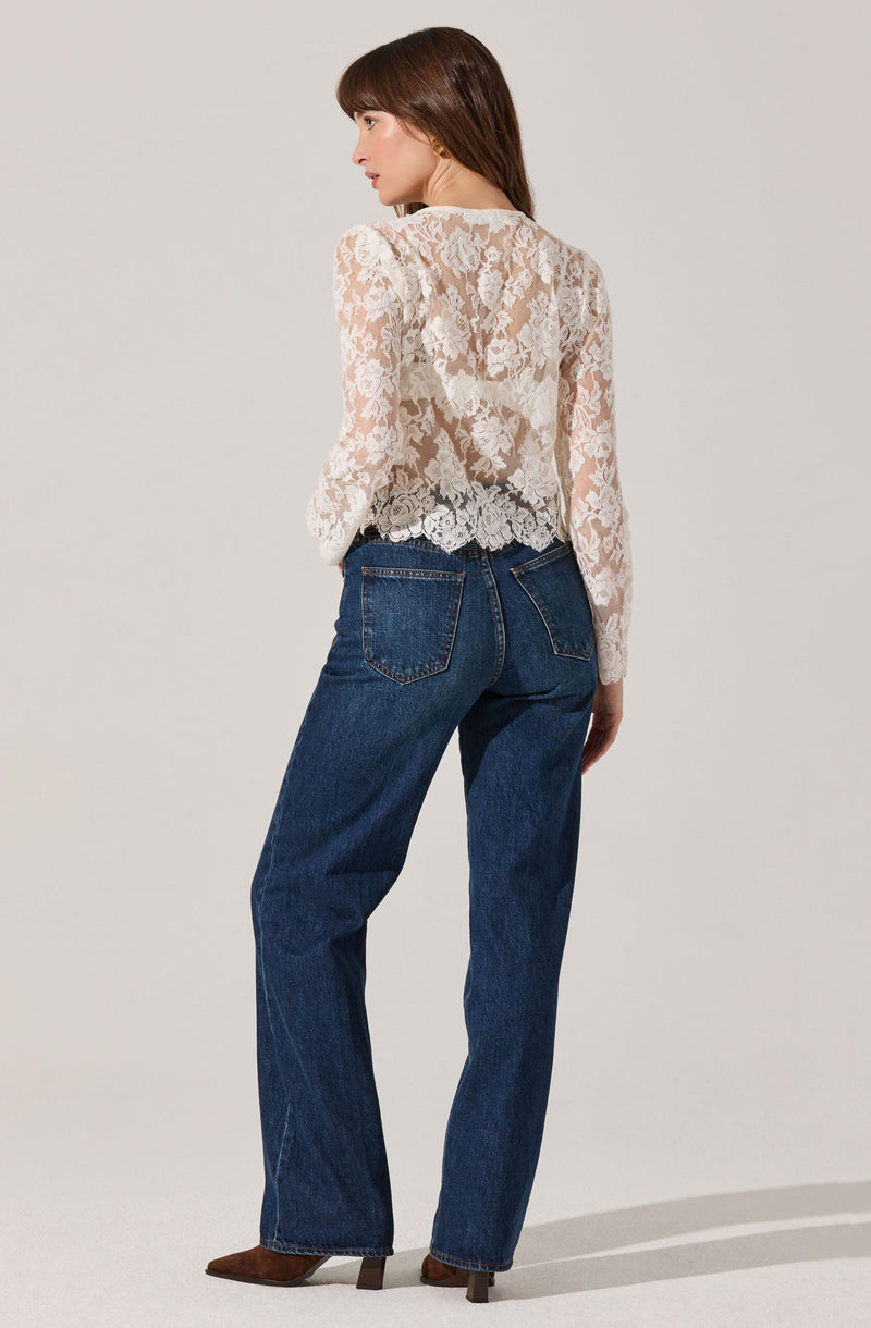 A woman is wearing a long-sleeved, white lace blouse with floral patterns, which is semi-transparent. She pairs it with high-waisted, dark blue jeans that have prominent stitching on the pockets and a relaxed fit. She is also wearing brown shoes with a pointed toe and a small heel.