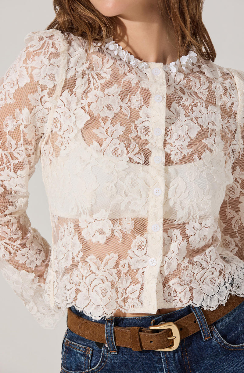 A woman is wearing a cream-colored lace button-up blouse with a floral pattern. The blouse features a round neckline adorned with small white floral embellishments, long puffed sleeves, and a scalloped hem. She pairs it with a brown belt and dark blue jeans.