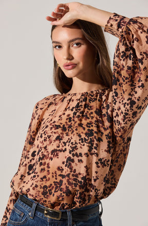 A woman is wearing a long-sleeved blouse with a round neckline, featuring a leopard print pattern in shades of brown, black, and beige. The blouse has a slightly loose fit and a gathered detail at the neckline. The fabric appears lightweight and silky. She has paired the blouse with dark blue high-waisted jeans and a black belt with a rectangular gold buckle.