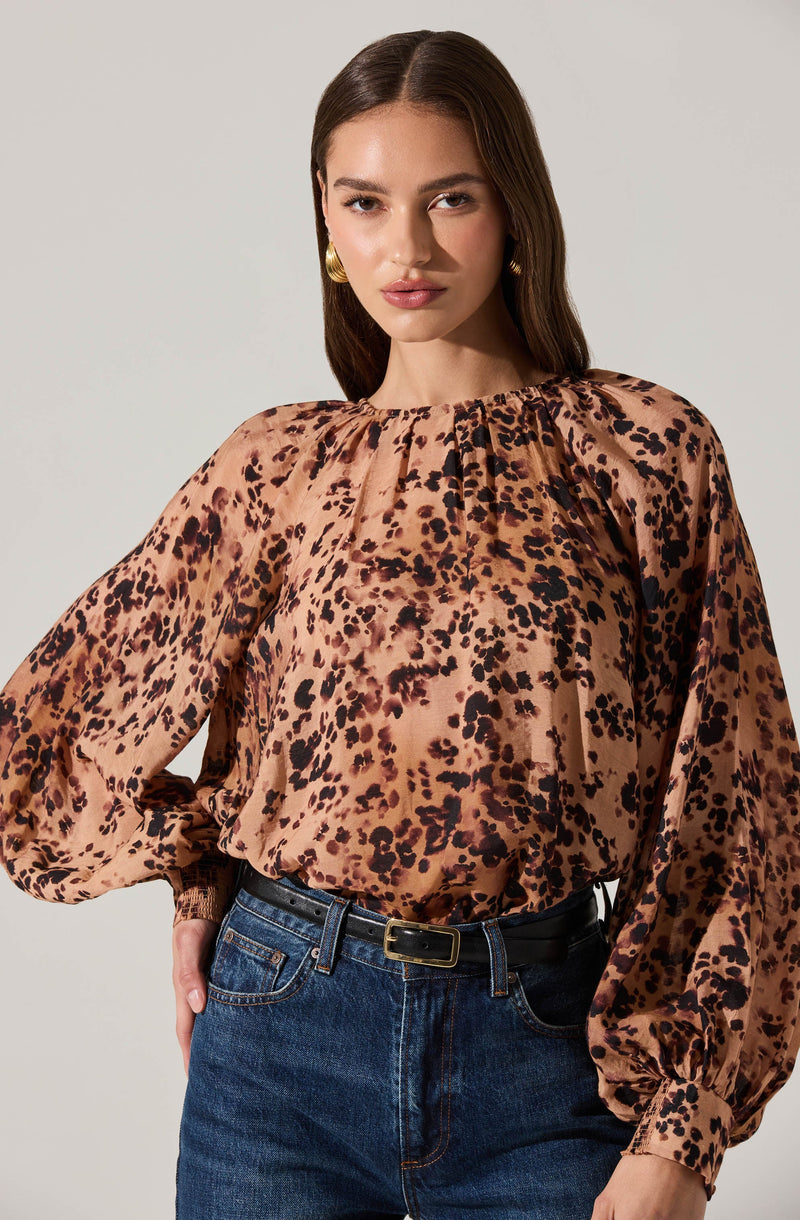 A woman is wearing a long-sleeved blouse with a leopard print pattern in shades of tan, brown, and black. The blouse has a gathered neckline with subtle pleats and voluminous sleeves that are cinched at the wrists. She is also wearing high-waisted blue jeans with a black belt.