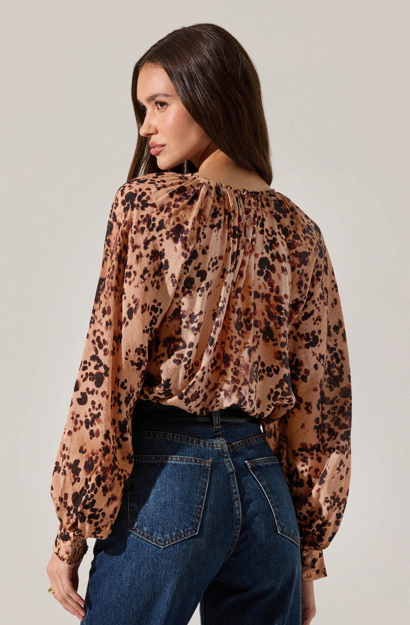 A woman is wearing a long-sleeved blouse with a leopard print pattern in shades of brown, black, and beige. The blouse features a gathered neckline with a small opening at the back, creating a slightly billowed effect on the sleeves and a relaxed fit. She pairs the blouse with high-waisted dark blue jeans.