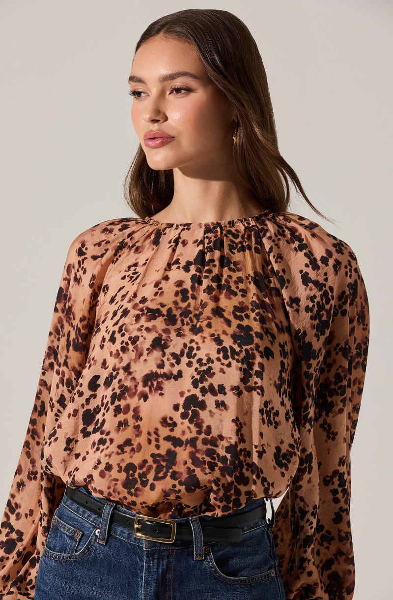 A woman is wearing a long-sleeved blouse with a leopard print pattern in shades of tan, brown, and black. The blouse has a gathered, round neckline and a loose, flowy fit. She pairs it with high-waisted blue jeans and a black belt with a gold buckle.