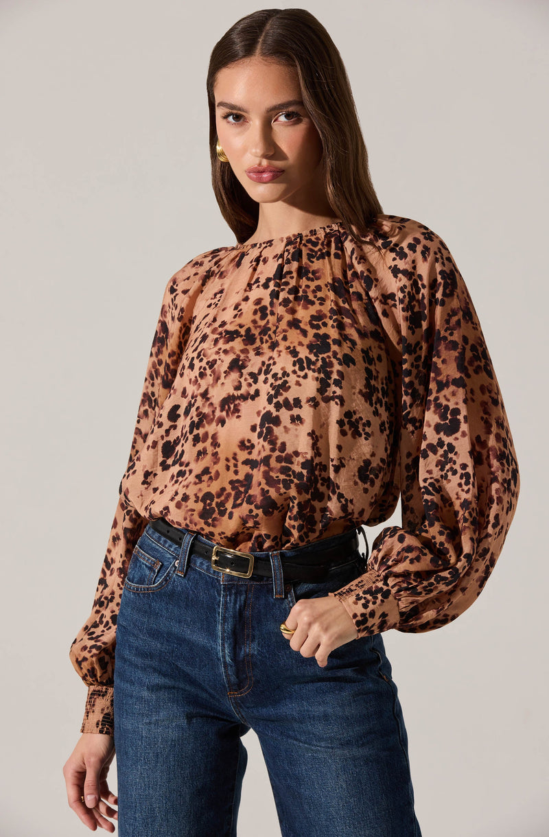 A woman is wearing a long-sleeved blouse with a rose gold and black leopard print pattern. The blouse has a gathered neckline, slightly puffed sleeves, and elasticized cuffs. She pairs it with high-waisted dark blue jeans and a black belt with a gold buckle. She has gold earrings and poses with one hand resting on her hip.