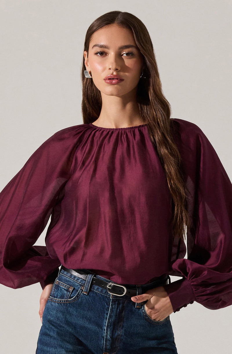 A woman is wearing a long-sleeved, deep burgundy satin blouse with a gathered neckline. The blouse has voluminous, slightly sheer sleeves that gather at the wrist with fitted cuffs. She pairs the blouse with high-waisted, dark blue jeans and a black belt with a silver buckle.
