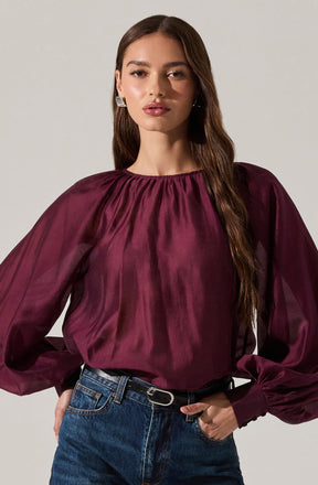 A woman is wearing a long-sleeved, deep burgundy satin blouse with a gathered neckline. The blouse has voluminous, slightly sheer sleeves that gather at the wrist with fitted cuffs. She pairs the blouse with high-waisted, dark blue jeans and a black belt with a silver buckle.