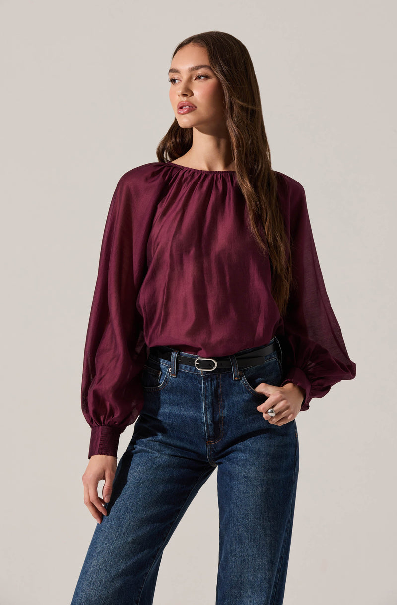 A woman is wearing a long-sleeved, satin-like blouse in a deep burgundy color with a gathered neckline and puffed sleeves that are fitted at the wrists. She pairs it with high-waisted, dark-wash jeans and a black belt with a silver buckle.