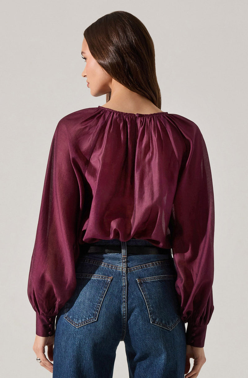 A woman is wearing a long-sleeved, shiny, deep burgundy satin blouse with a gathered neckline and slightly puffed sleeves that are cuffed at the wrists. She pairs it with high-waisted blue jeans.