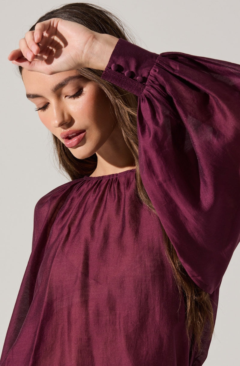 A woman is wearing a loose-fitting, long-sleeved blouse made of shiny, satin-like fabric in a deep burgundy color. The blouse features gathered fabric at the neckline and wide, puffed sleeves with buttoned cuffs.