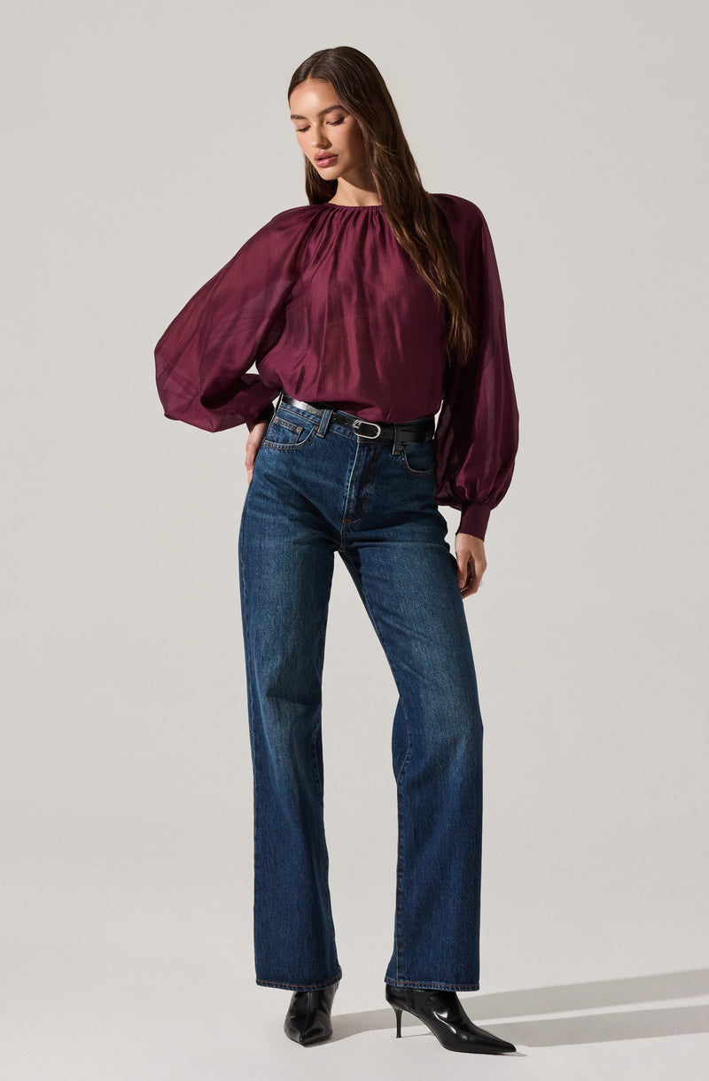 A woman is wearing a long-sleeved, burgundy satin blouse with a gathered neckline and voluminous sleeves that taper at the wrists. She pairs the blouse with high-waisted, dark-wash straight-leg jeans and black pointed-toe high-heeled boots.